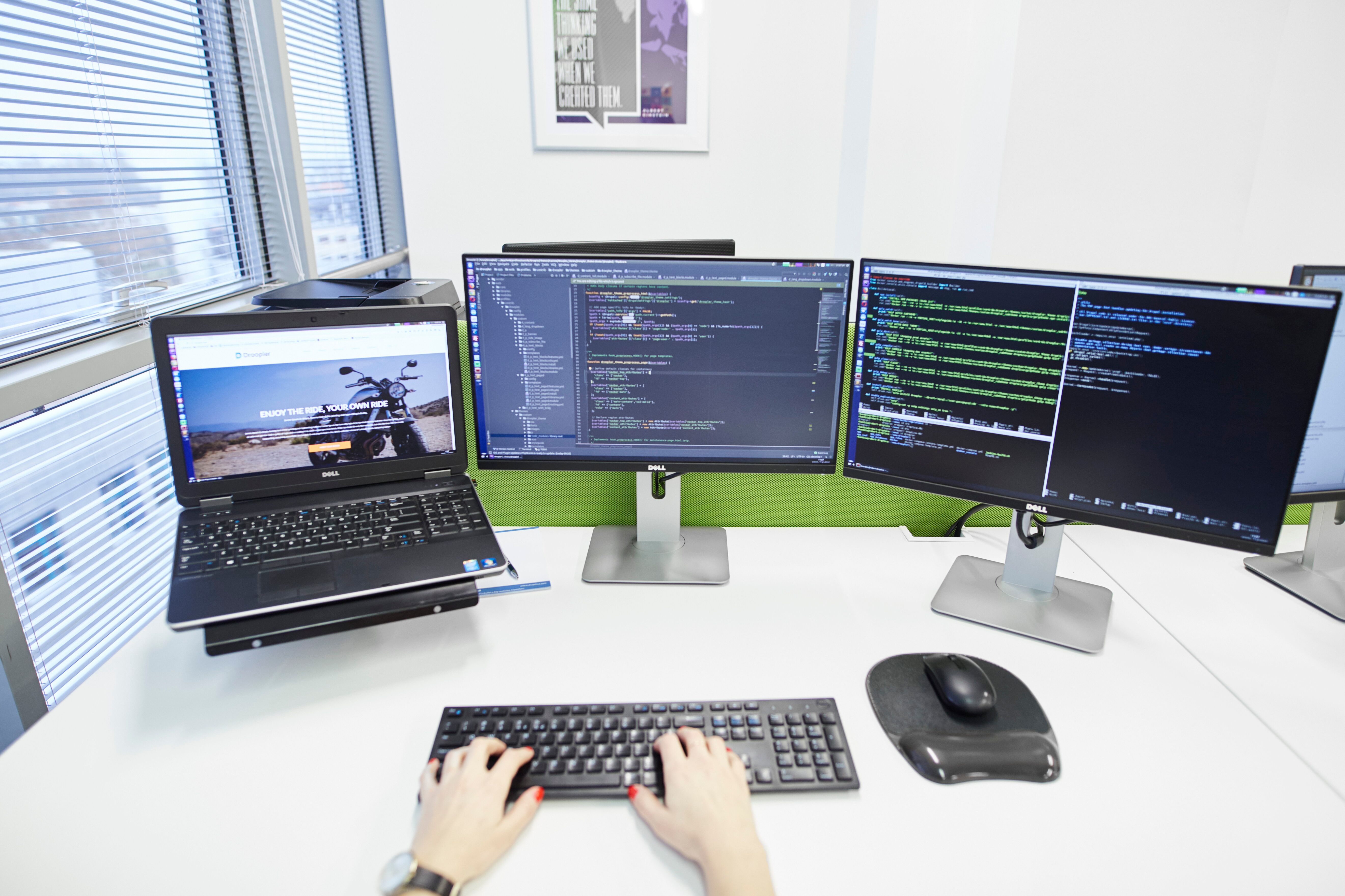 Zdjęcie stanowiska pracy. Widoczne są dwa monitory, mysz na podkładce i klawiatura, na której oparte są czyjeś dłonie